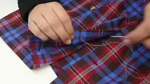 Knopf annähen: Mit der Nadel wieder zur Oberseite des Hemdes durchstechen, den Knopf einfädeln und mittels Kreuzstich (abwechselnd diagonal in die Löcher des Knopfes einstechen) den Knopf annähen. Diesen Vorgang zweimal wiederholen.